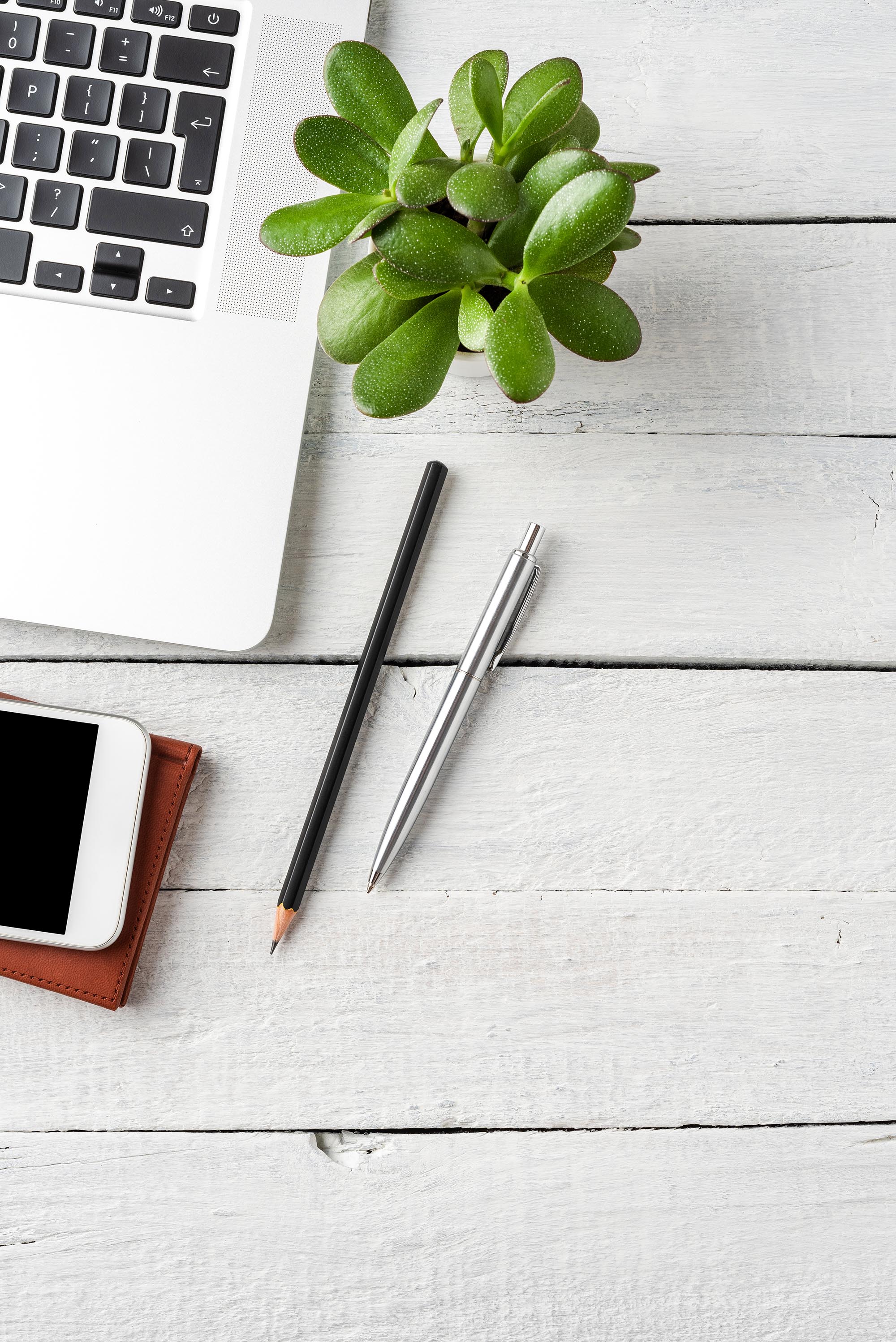 Laptop, mobile phone, notebook, pen and small flower on white wooden desktop